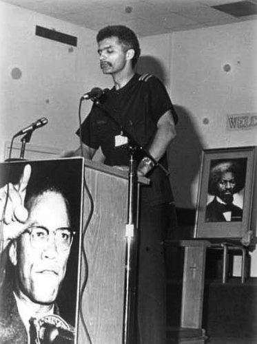 Tall thin New Afrikan man at podium with posters of Malcolm X and Frederick Douglass