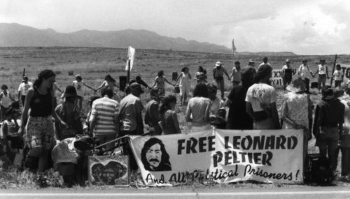 Photo of dozens of people by a roadside holding protest signs including Free Leonard Peltier