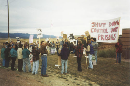 A couple dozen people in jackets and jeans hold placards by the entrance road to the prison