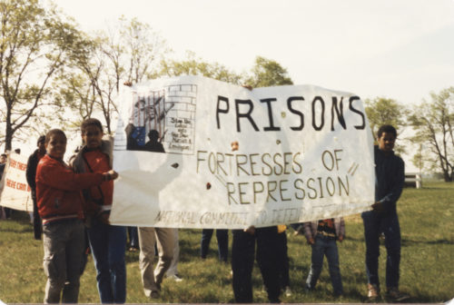 Half a dozen demonstrators standing on grassy field and holding banner "Prisons Are Fortresses of Repression"