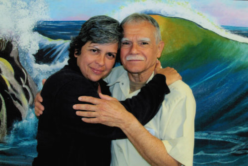 Photo of adult woman with dark hair and serious smile embracing older man with gray hair and mustache